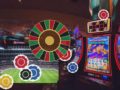 Casino floor with slot machines on the right and a large colorful roulette wheel, overlaid with chip icons in red, blue, yellow, and black.